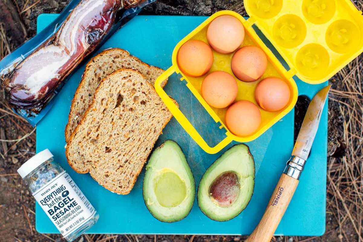 Ingredientes para hacer un sándwich de desayuno con tostadas de aguacate (tocino, huevos, aguacate, pan) sobre una tabla de cortar azul