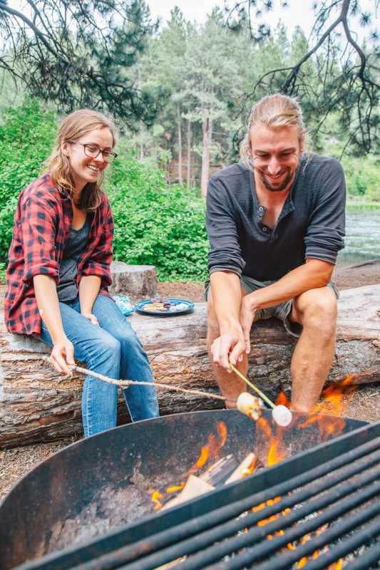 Megan e Michael sono seduti su un tronco e arrostiscono marshmallow su un fuoco da campo