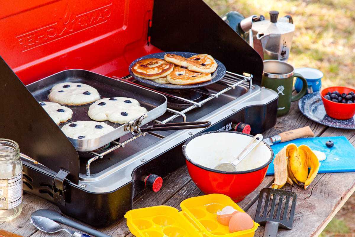 キャンプ用ストーブの上の鉄板に置かれたパンケーキと、さまざまなキャンプ用調理器具がフレームに入っています