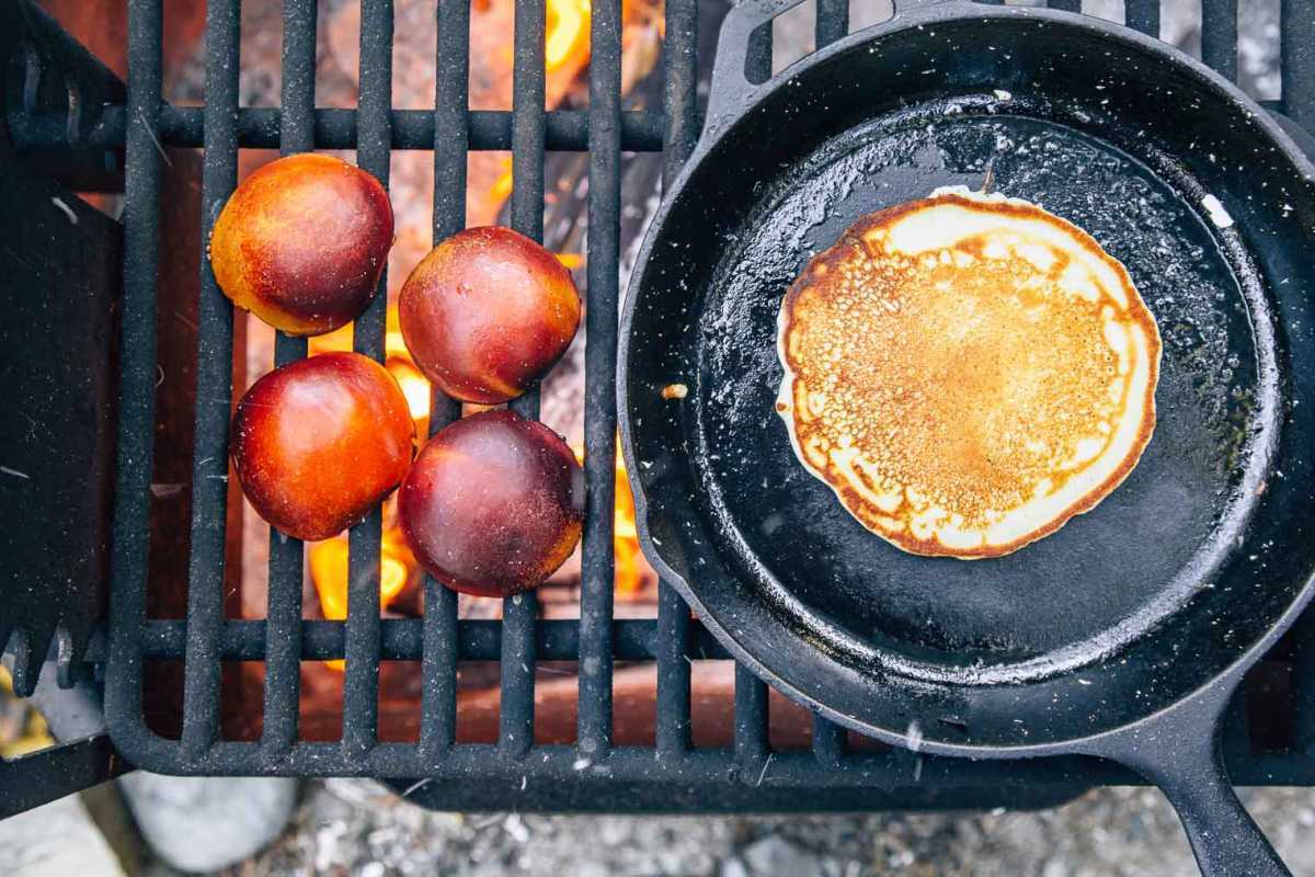 Pancake dalam wajan dan buah persik yang dibelah dua di atas panggangan api unggun