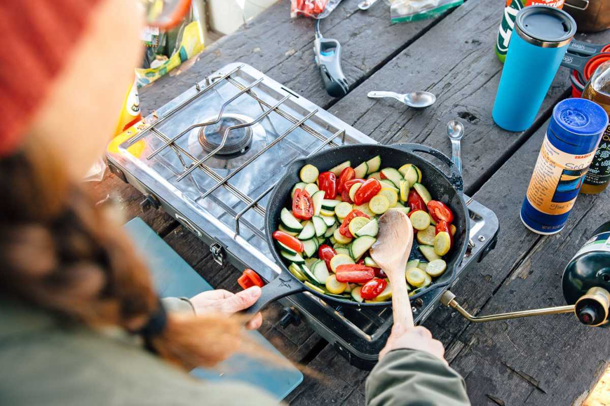 מטגנים עגבניות, זוקיני ודלעת קיץ צהובה במחבת ברזל יצוק על כיריים קמפינג.