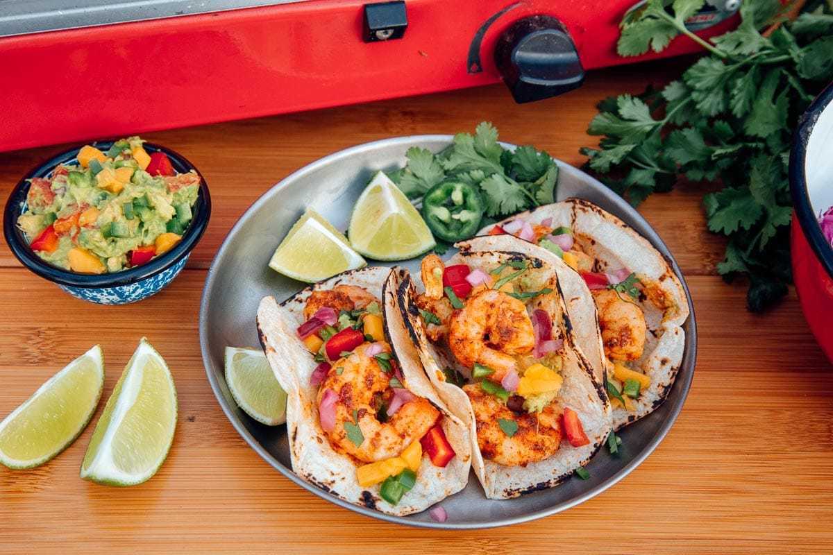 Tri krevetové tacos na striebornom tanieri s nakrájanými limetkami a malou miskou guacamole na boku.