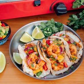 Trzy tacos z krewetkami na srebrnym talerzu z plasterkami limonki i małą miską guacamole na boku.