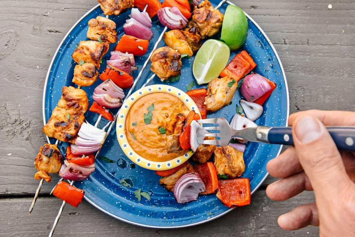 Brochettes de poulet grillées thaïlandaises sur une assiette de camping bleue avec un petit bol de trempette aux arachides.