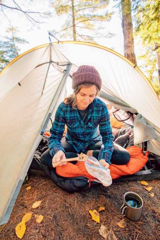 Megana sēž pie mugursomas telts ieejas un ēd granolu no somas
