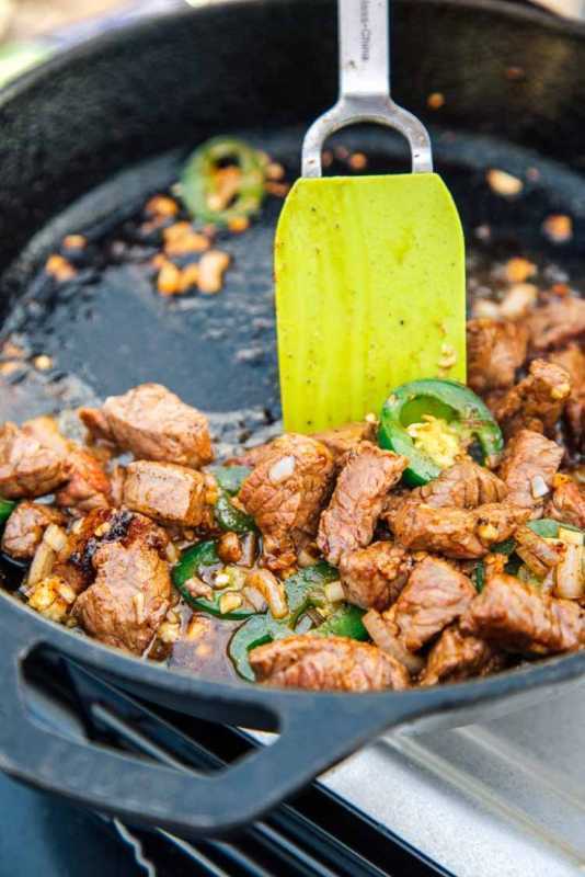 Bidikan aksi dari spatula yang menumis steak, jalapeños, bawang putih, dan bawang merah dalam wajan besi cor.