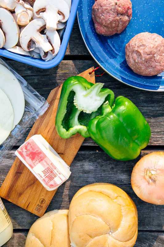 Ingredientes em uma mesa, incluindo pimentão verde, cebola, pães e carne de hambúrguer.