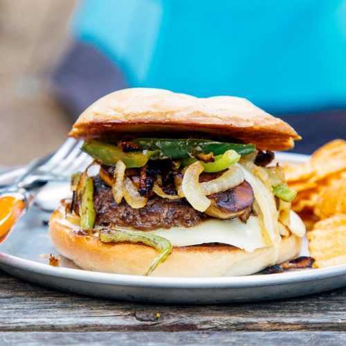 En burger toppet med grønn paprika og løk på en blå tallerken og campingscene i bakgrunnen