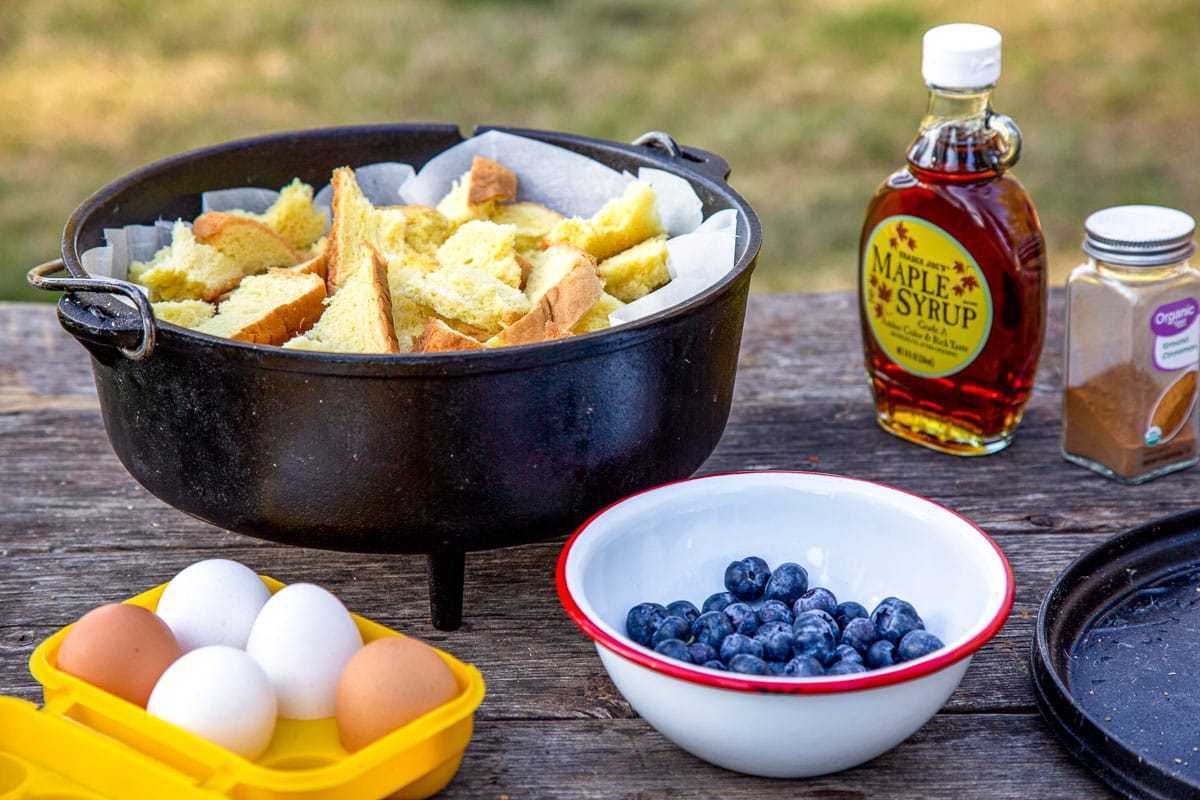테이블 위에는 찢어진 빵, 블루베리 한 그릇, 계란이 담긴 노란색 달걀 상자, 메이플 시럽 한 병이 가득 담긴 더치 오븐.