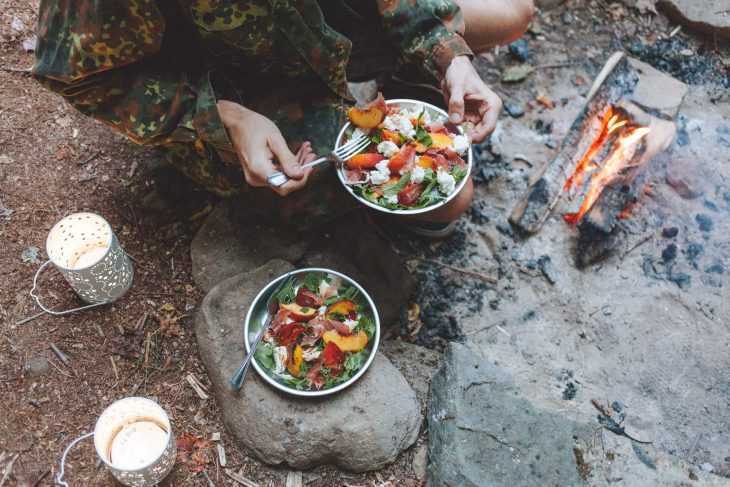 Persoană care ține o farfurie cu salată caprese de piersici lângă un foc de tabără